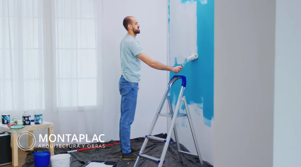 Hombre pintando una pared azul con rodillo durante una reforma interior; trabajo doméstico sencillo representativo de una obra menor en vivienda.